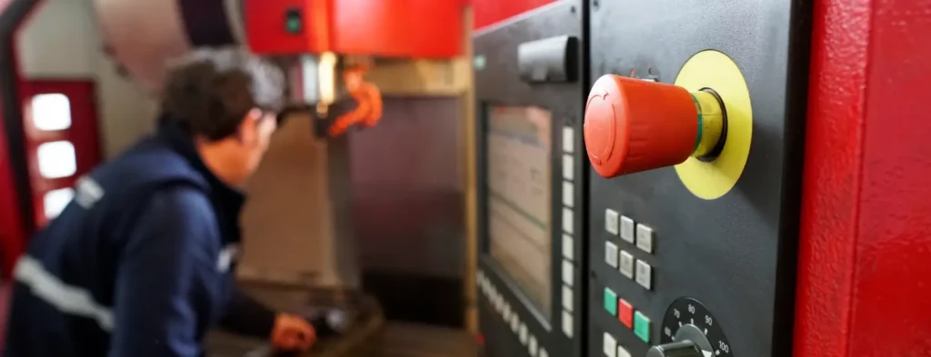 CNC machine operator inspecting equipment beside a Fanuc control panel with emergency stop button in the foreground