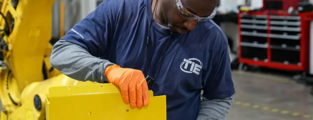 Technician wearing safety glasses and orange gloves servicing a yellow industrial robot component in a workshop